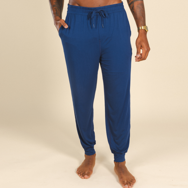 Barefoot against a cream wall in navy MicroModal pajama pants, tattooed hand in the pocket, gold watch on