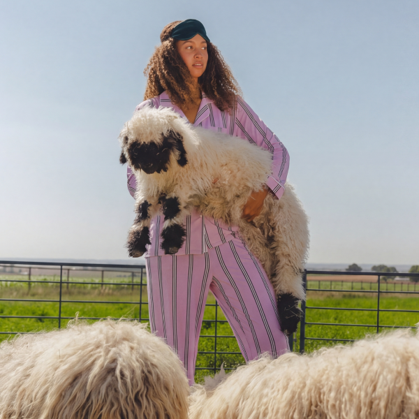 Woman in lavender striped pajamas and a sleep mask carrying a black-nosed sheep through a pasture