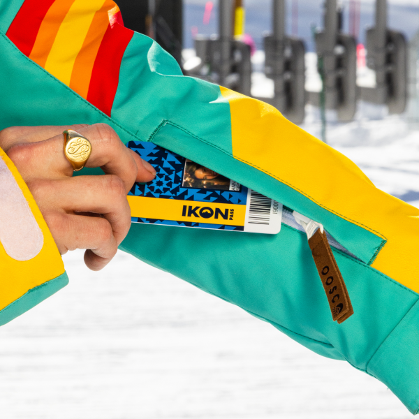 Hand pulling a lift pass from the sleeve pocket of a teal Shinesty ski suit, lift towers behind