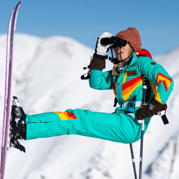 Skier in a teal retro one-piece Shinesty ski suit with rainbow stripes kicking a leg out on a snowy ridge